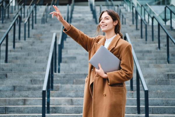 Young smiling businesswoman in coat with laptop joyfully waving
